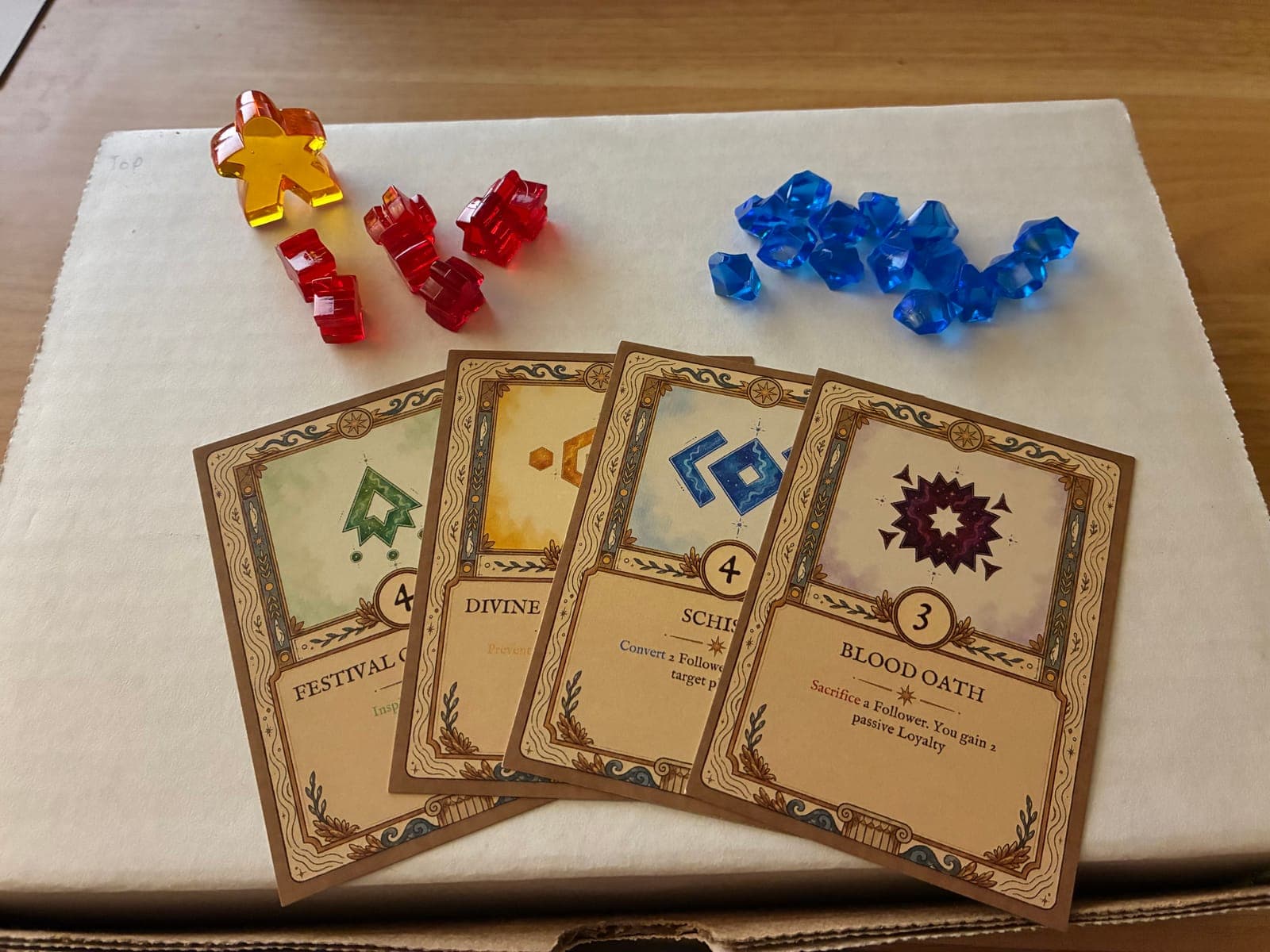 Printed prototype cards — Festival, Divine, Schism, and Blood Oath — laid out beside red and blue resource gems and a meeple