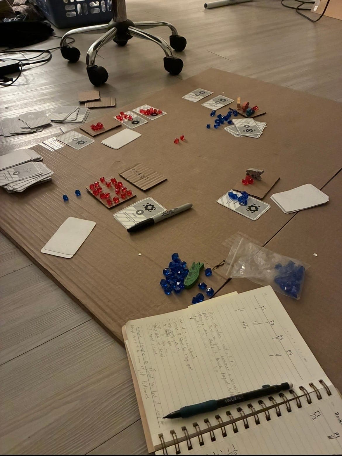 A playtest in progress on a cardboard board, with player areas, gem stockpiles, and a notebook of balance notes